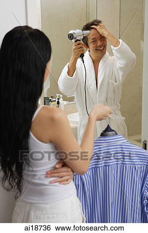 Stock Photograph - Couple in bathroom, man in robe using hairdryer, woman holding shirt on hanger. Fotosearch
