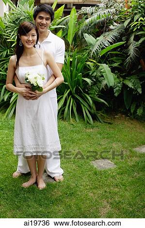 Stock Photograph - Couple in garden, man with arms around woman, woman holding bouquet. Fotosearch