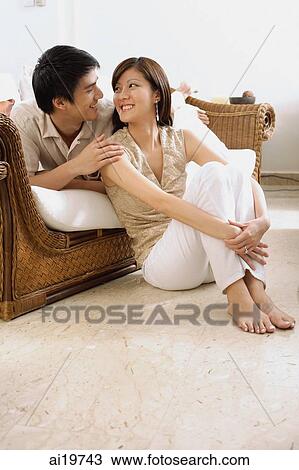 Stock Image - Couple in living room, smiling at each other. Fotosearch