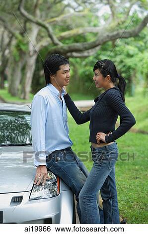 Stock Photograph - Couple looking at each other, man leaning on hood of car. Fotosearch