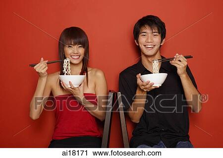 Couple sitting against red wall, holding bowl of noodles, smiling at camera View Large Photo Image Stock Image - Couple sitting against red wall, holding bowl of noodles, smiling at camera. Fotosearch