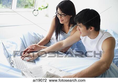 Couple sitting on bed using laptop View Large Photo Image Stock Image - Couple sitting on bed using laptop. Fotosearch