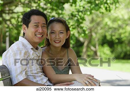 Couple sitting on bench, looking at camera View Large Photo Image Stock Photography - Couple sitting on bench, looking at camera. Fotosearch