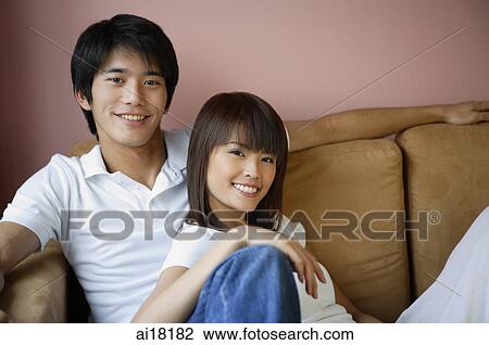 Stock Image - Couple sitting on sofa, smiling at camera. Fotosearch