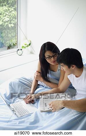 Stock Image - Couple sitting side by side in bedroom, using laptop. Fotosearch
