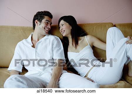 Stock Image - Couple sitting side by side on sofa, looking at each other. Fotosearch