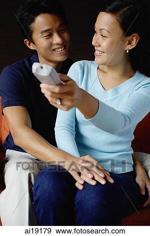 Stock Photo - Couple sitting side by side, woman holding TV remote control. Fotosearch