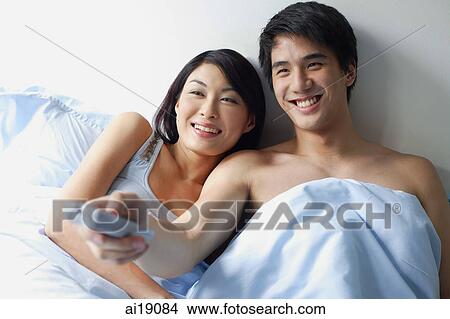 Couple sitting up in bed, watching TV, man holding TV remote control View Large Photo Image Picture - Couple sitting up in bed, watching TV, man holding TV remote control. Fotosearch