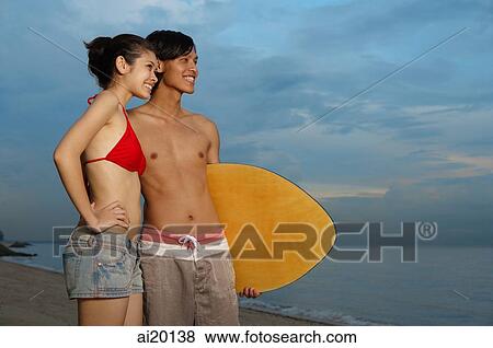 Stock Photo - Couple standing on beach, man carrying skimboard, looking away. Fotosearch