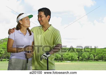 Stock Image - Couple standing side by side, holding golf clubs, looking at each other. Fotosearch