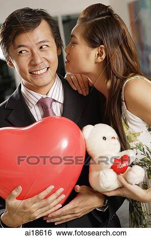Couple standing side by side, man holding heart shaped balloon, woman holding teddy bear, kissing man on cheek View Large Photo Image Stock Photograph - Couple standing side by side, man holding heart shaped balloon, woman holding teddy bear, kissing man on cheek. Fotosearch