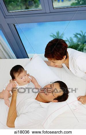 Father lying on bed, holding baby girl, mother looking on View Large Photo Image Stock Photograph - Father lying on bed, holding baby girl, mother looking on. Fotosearch
