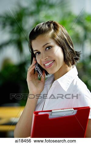 Stock Photograph - Female executive carrying clipboard, using mobile phone, smiling. Fotosearch