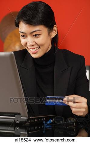 Picture - Female executive sitting at desk, in front of laptop, holding credit card. Fotosearch