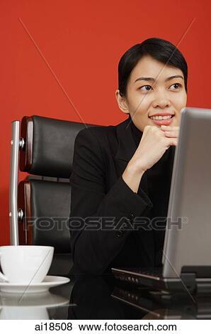 Stock Photo - Female executive sitting at desk, in front of laptop, hand on chin, looking away. Fotosearch