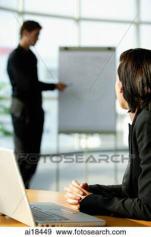 Stock Photo - Female executive sitting at desk, turning to look at colleague standing next to flip board. Fotosearch
