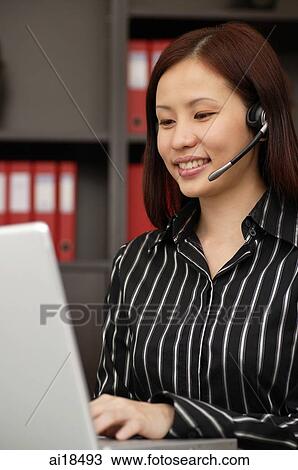 Stock Image - Female executive wearing hands free device, using laptop. Fotosearch