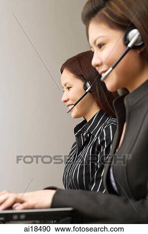 Stock Image - Female executives wearing hands free devices, using laptops. Fotosearch