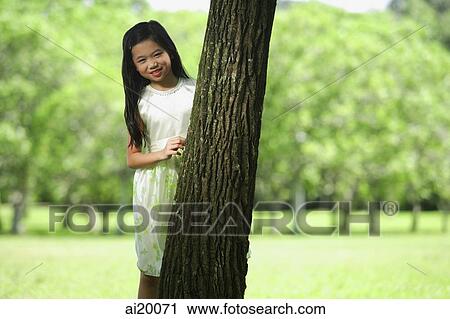 Stock Image - Girl standing behind tree trunk, smiling at camera. Fotosearch