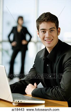 Male executive dressed in black, sitting next to laptop, looking at camera, smiling View Large Photo Image Stock Image - Male executive dressed in black, sitting next to laptop, looking at camera, smiling. Fotosearch