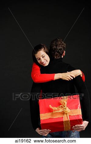Stock Image - Man holding gift behind his box, woman embracing him. Fotosearch