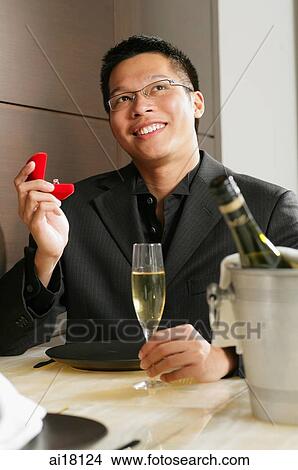 Picture - Man holding ring box and glass of champagne, smiling, looking away. Fotosearch