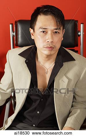 Stock Image - Man in black shirt and beige jacket, sitting on chair, looking at camera. Fotosearch