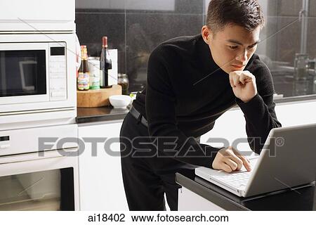 Stock Image - Man in kitchen, looking at laptop. Fotosearch