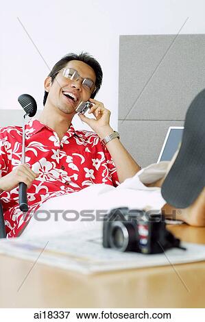 Stock Photo - Man in printed shirt sitting in office, using mobile phone, feet up on desk. Fotosearch