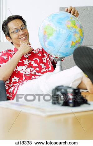 Stock Photo - Man in printed shirt sitting in office, feet up on desk, looking at globe. Fotosearch