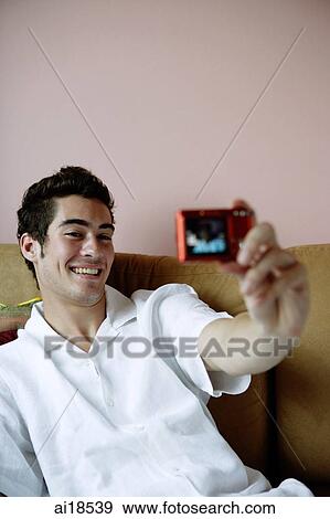 Stock Photo - Man on sofa, holding camera, taking photograph of himself. Fotosearch