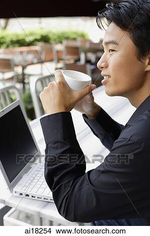 Man sitting in front of laptop, holding cup, looking away View Large Photo Image Picture - Man sitting in front of laptop, holding cup, looking away. Fotosearch