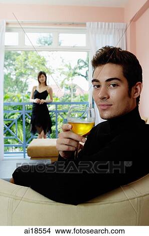 Picture - Man sitting in living room, holding a glass of wine, woman in the background. Fotosearch