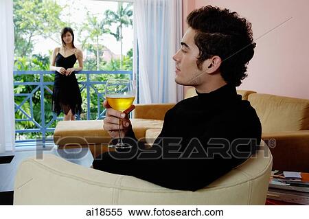Stock Photography - Man sitting in living room with a glass of wine, woman standing in the balcony. Fotosearch
