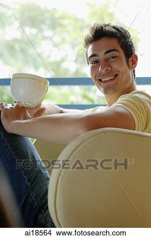 Man sitting on armchair, holding cup, smiling at camera View Large Photo Image Picture - Man sitting on armchair, holding cup, smiling at camera. Fotosearch