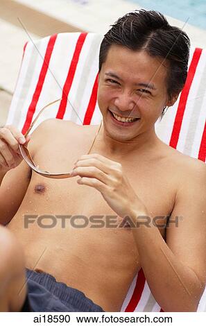 Stock Image - Man sitting on chair by swimming pool, holding sunglasses, smiling. Fotosearch