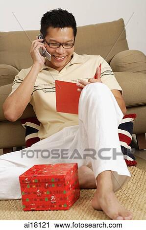 Stock Image - Man sitting on floor, looking at card, using mobile phone. Fotosearch