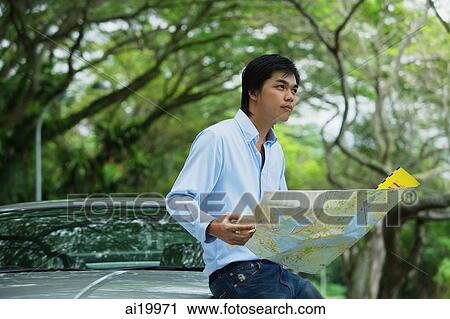 Stock Image - Man sitting on hood of car, holding map, looking away. Fotosearch