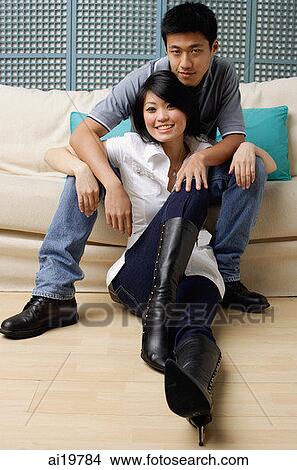 Man sitting on sofa, woman sitting on floor, smiling at camera View Large Photo Image Picture - Man sitting on sofa, woman sitting on floor, smiling at camera. Fotosearch