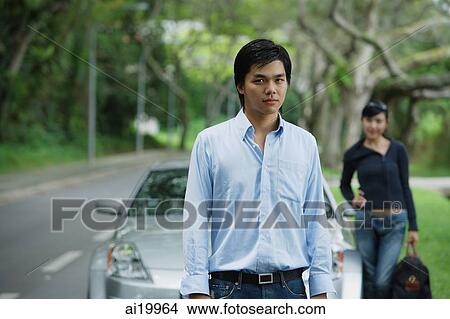 Picture - Man standing in front of car, woman in the background with luggage. Fotosearch