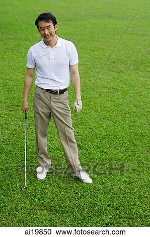 Stock Image - Man standing on golf course, holding golf club, smiling. Fotosearch