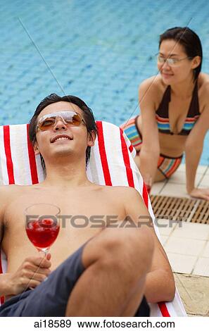 Stock Photo - Man sun tanning, holding wine glass, woman sitting at edge of swimming pool behind him. Fotosearch