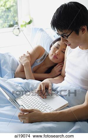 Picture - Man using laptop, woman next to him on bed, sleeping. Fotosearch