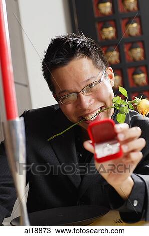 Stock Image - Man with rose stalk between teeth, holding ring box towards camera, smiling. Fotosearch