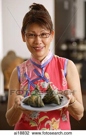 Stock Image - Mature woman holding plate of dumplings, smiling at camera. Fotosearch