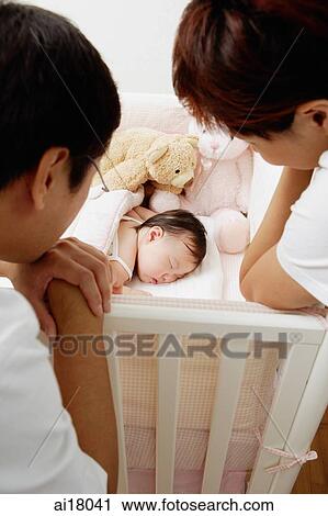 Stock Image - Mother and father leaning on crib looking down at sleeping baby. Fotosearch