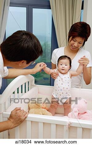 Picture - Mother helping baby girl stand in crib, father holding her hand. Fotosearch