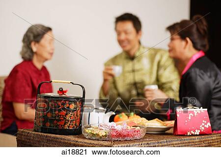 Stock Image - People having tea in living room, traditional Chinese New Year goodies in the foreground. Fotosearch