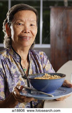 Stock Photograph - Senior woman holding bowl of noodles on a plate, looking at camera. Fotosearch