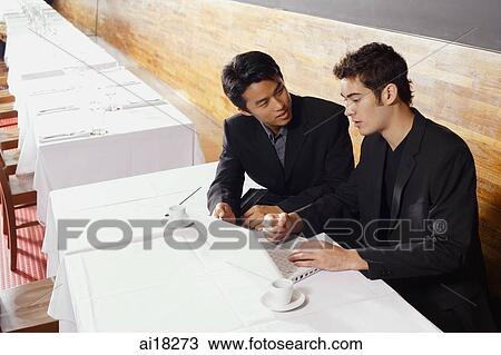Stock Image - Two men sitting at table in restaurant, using laptop. Fotosearch